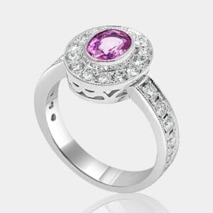 Handmade, designer style engagement ring featuring oval cut pink sapphire and halo of round diamonds, with diamonds in the band and milgrain detail.