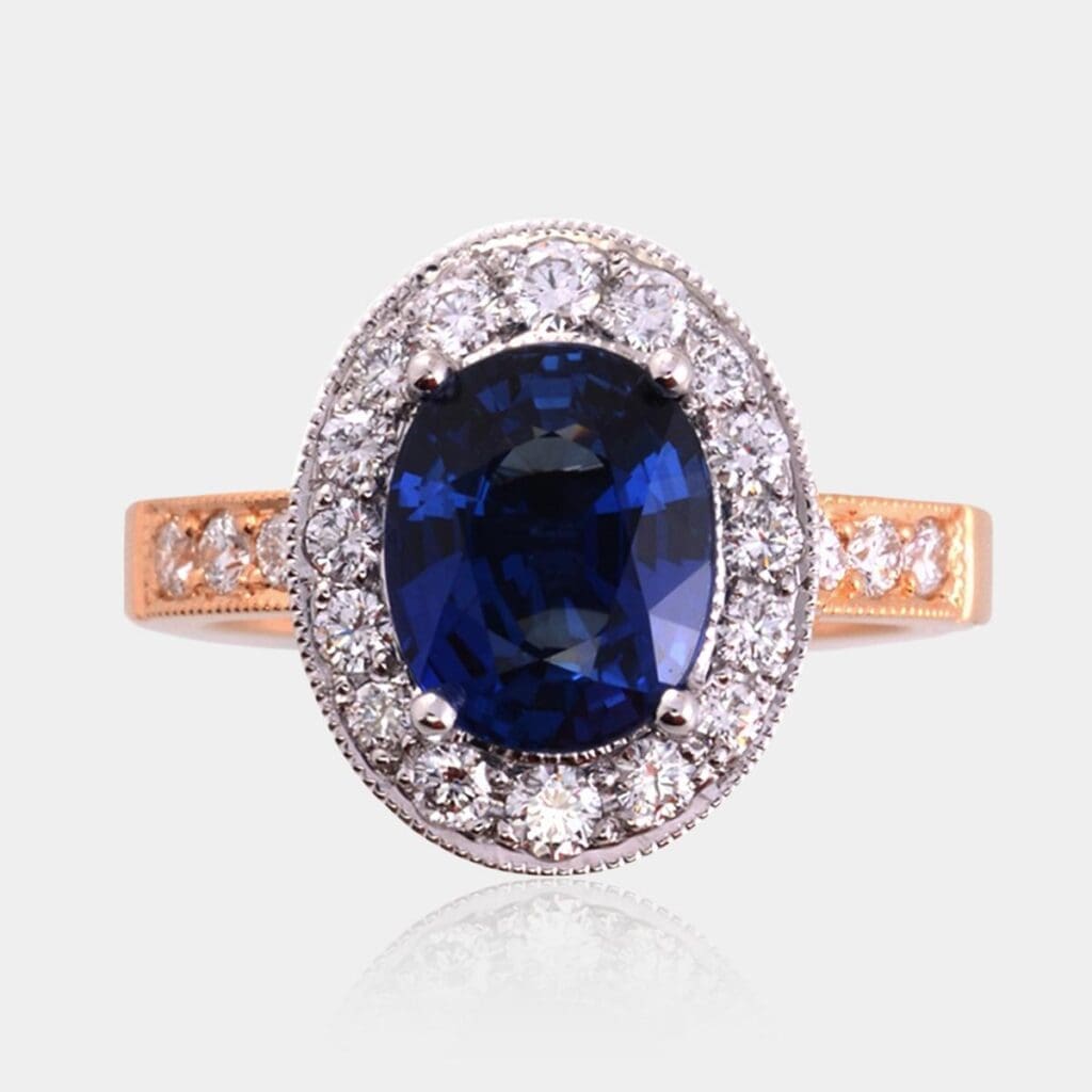 Halo style engagement ring set with oval cut sapphire surrounded by round brilliant cut diamonds in white gold. Yellow gold band bead set with round diamonds and milgrain finish.